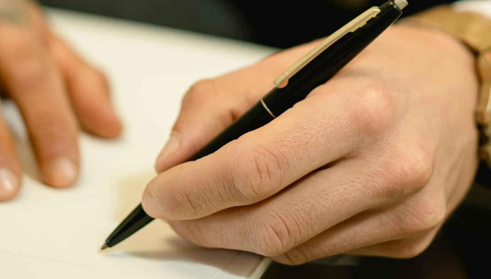 Primer plano de una mano firmando un documento con un bolígrafo, sobre un escritorio claro.